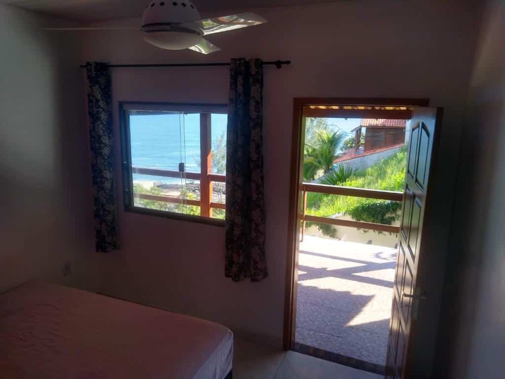 Quarto do Farol House. Uma cama de casal do lado esquerdo, de frente uma janela e do lado direito a porta do quarto. Uma varanda com vista para o mar. Foto para ilustrar post sobre pousadas em Maricá.