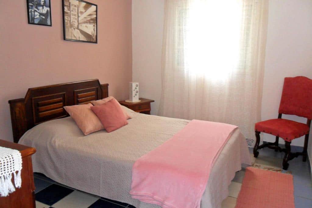 Foto do quarto da Fazenda Monte Verde A Morada do Muriqui. Representa o post sobre airbnb em São Francisco Xavier. Há uma cama de casa na esquerda. Na direita da cama há uma janela com cortinas e uma cadeira vermelha.