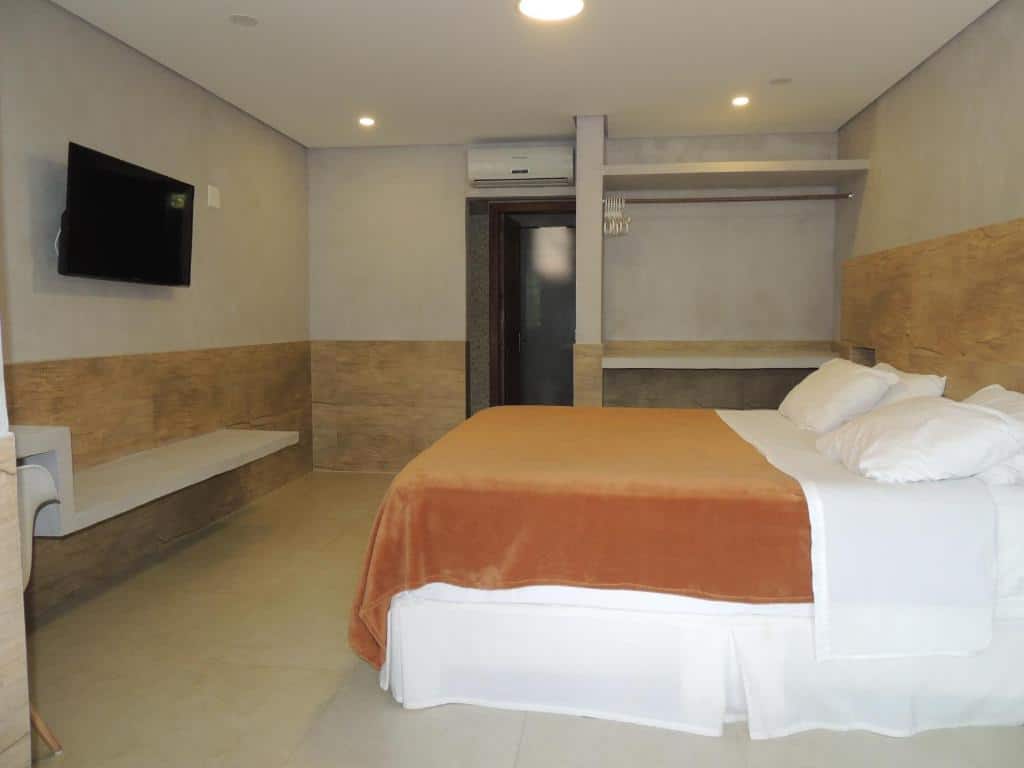 Quarto espaçoso do Flat Camburi, um dos airbnb em Camburi. Uma cama de casal com cobertor, jogo de cama e travesseiros está encostada na parede ao lado direito e encara uma mesinha com cadeira na parede oposta, onde há uma TV logo acima. Ao lado da cama há um espaço com cabides e um aparelho de ar-condicionado.