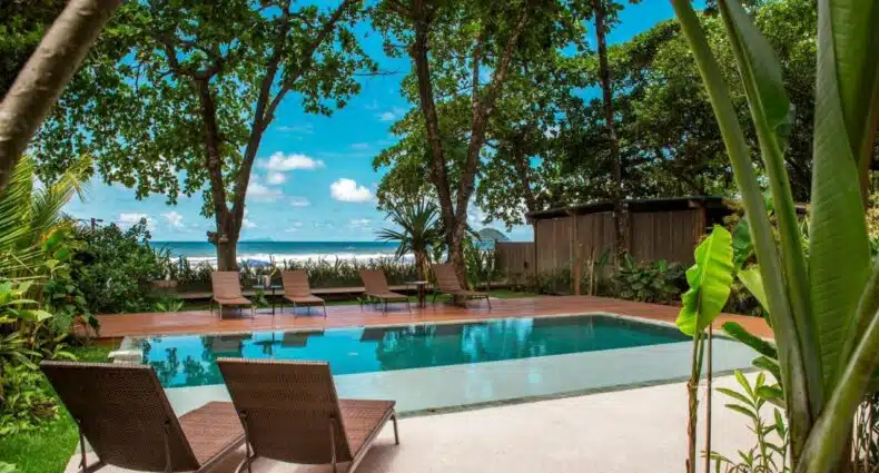 Espreguiçadeiras encarando a piscina cercada por plantas no Flat Pitanga, um dos airbnb em Camburi. Ao fundo é possível ver um pouco da praia. O céu acima é azul com poucas nuvens.