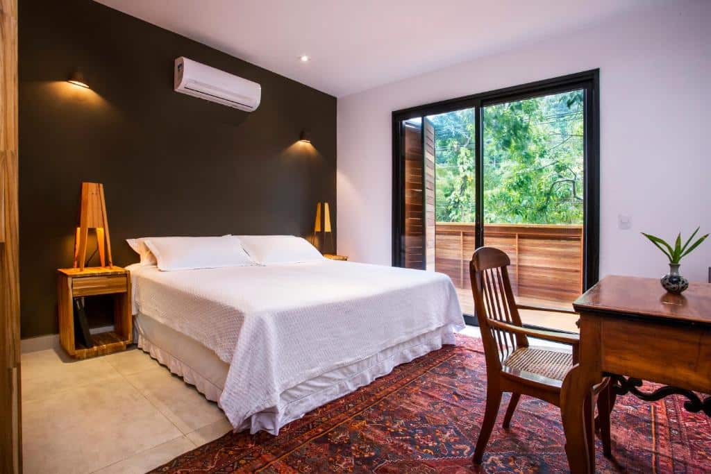 Quarto do Flat Pitanga, um dos airbnb em Camburi. Uma cama de casal com travesseiros e jogo de cama está encostada na parede ao lado esquerdo e tem mesinhas de cabeceira com abajur dos dois lados, assim como um aparelho de ar-condicionado na parede acima. Ela está de frente para uma mesa de trabalho com cadeira, que fica em cima de um tapete. Na parede ao lado direito da cama fica uma porta de vidro com vista para a varanda.