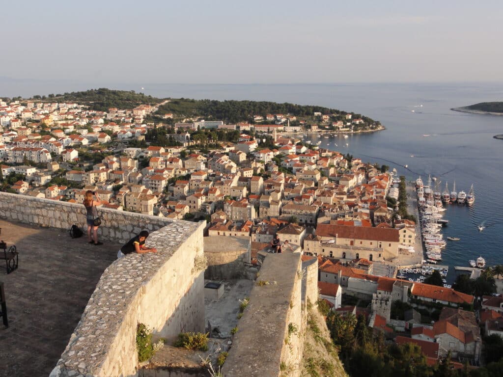 Vista aérea de Hvar no Forte Espanhol com edifícios com telhados vermelhos, um porto com vários barcos e um muro de pedra em primeiro plano.