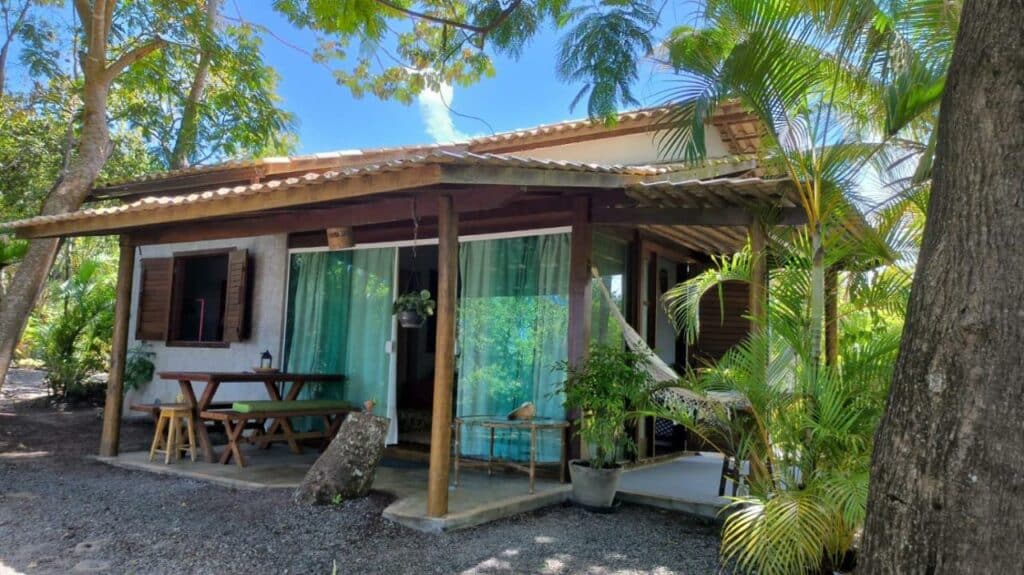 Frente do  Chalé "Oasis" durante o dia com a estadia na frente ao fundo do lado esquerdo na varada uma mesa com bancos. Representa airbnb na Costa do Sauípe
