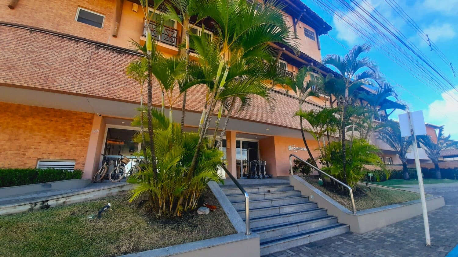 Fachada do Hotel Ponta Verde Francês, contendo uma escada com jardins nos dois lados, e o prédio do hotel possui portas de vidro no térreo e quartos com sacadas nos andares superiores