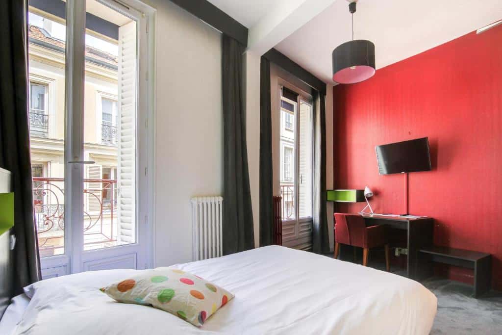 Quarto do Hôtel d'Angleterre, um dos hotéis perto do Palácio de Versalhes. Uma cama de casal encostada na parede esquerda encara uma mesa com cadeira, abajur e TV acima na parede oposta. Na lateral esquerda há duas janelas com cortinas.
