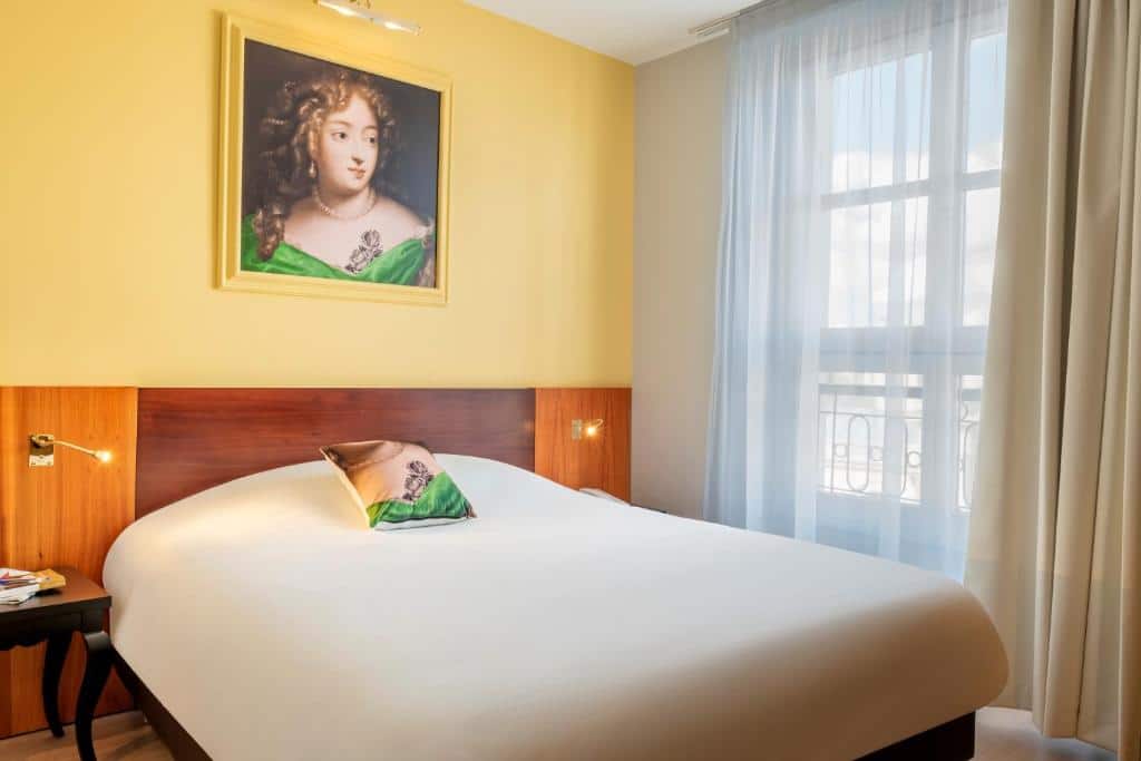 Quarto do Hotel des Lys, um dos hotéis perto do Palácio de Versalhes. Uma cama de casal com mesinhas de cabeceira e luminárias dos dois lados está encostada na parede ao lado esquerdo e tem uma janela com cortinas logo ao lado. Na parede acima da cama está um quadro.
