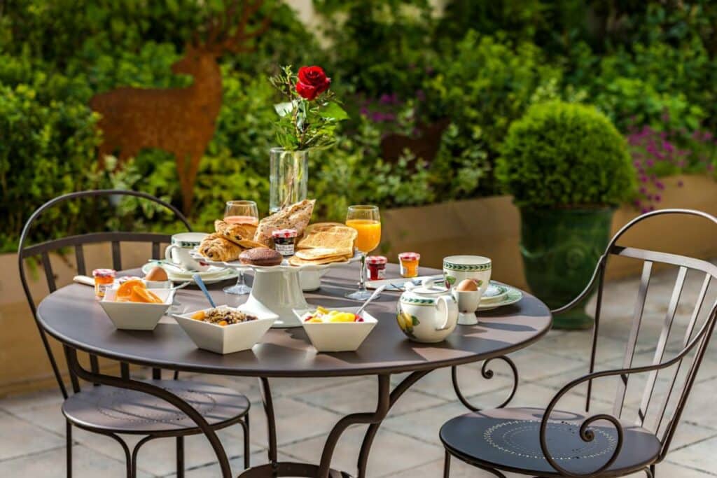 Mesa de café da manhã no jardim do Hôtel Le Versailles com duas cadeiras. Em cima da mesa há uma rosa e taambém diversos itens comestíveis, como frutas, pães, frios, ovos, geléias, taças de suco e xícaras de café. Ao fundo estão algumas plantas e flores.