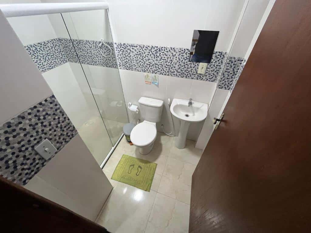 Banheiro do Hotel Marquês de Maricá. Um vaso sanitário, uma pia, um espelho, papel higiênico e lixo do lado direito, do lado esquerdo o boz de vidro.