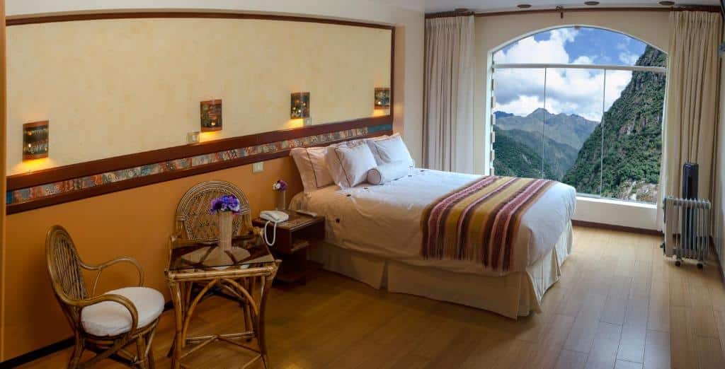 quarto do Hotel Taypikala Machupicchu com ampla janela que dá vista para as montanhas, uma cama de casal e uma mesa pequena para dois lugares ao lado.