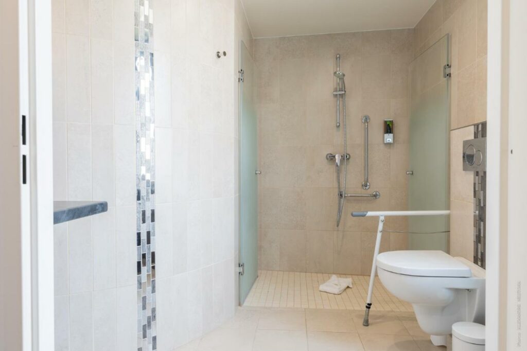 Banheiro adaptado para PCD no Hôtel Versailles Chantiers. Na parede ao lado direito está aprivada e a lixeira ao lado do box com chuveiro. Há barras de apoio ao lado da privada e na área de banho.