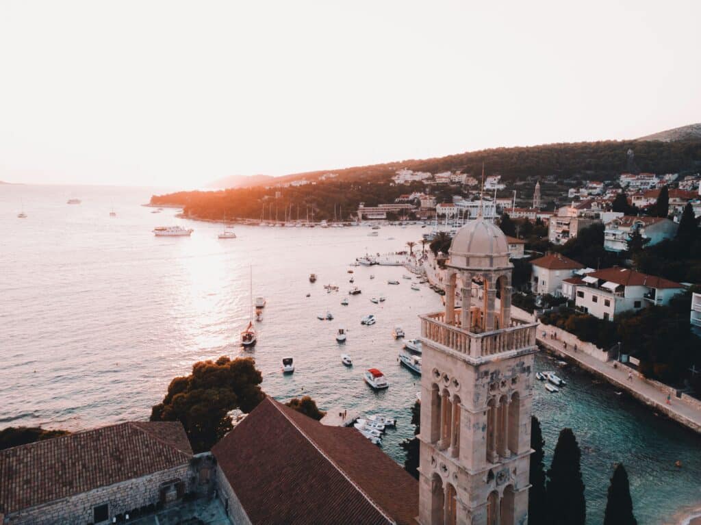 Vista aérea de Hvar na Croácia, com uma torre de igreja proeminente, barcos na água e colinas ao fundo durante o pôr do sol.