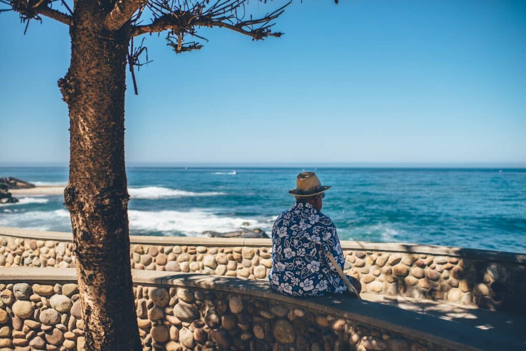 Um senhor usando um chapéu sentada em um banco de frente para um mar de água azul clara, próximo dele também há uma árvore, para representar seguro viagem para idosos