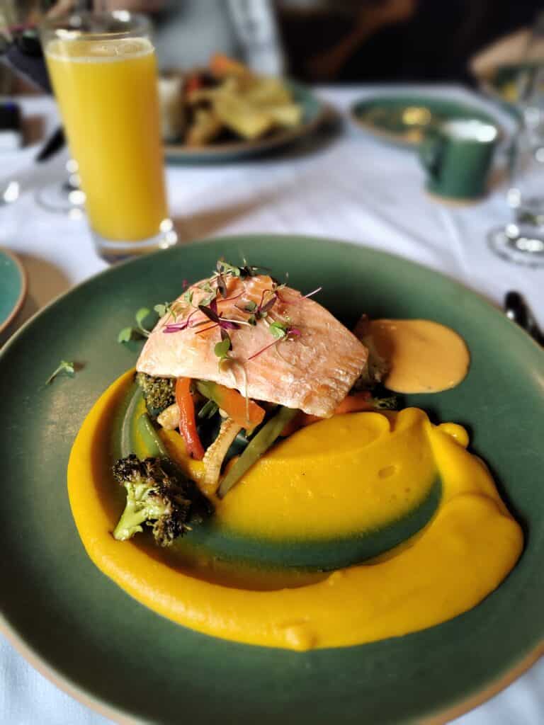 prato circular verde com um pedaço de truta grelhada e purê de abóbora bem amarelo embaixo, junto com alguns legumes, como brócolis, cenoura e pimentões.