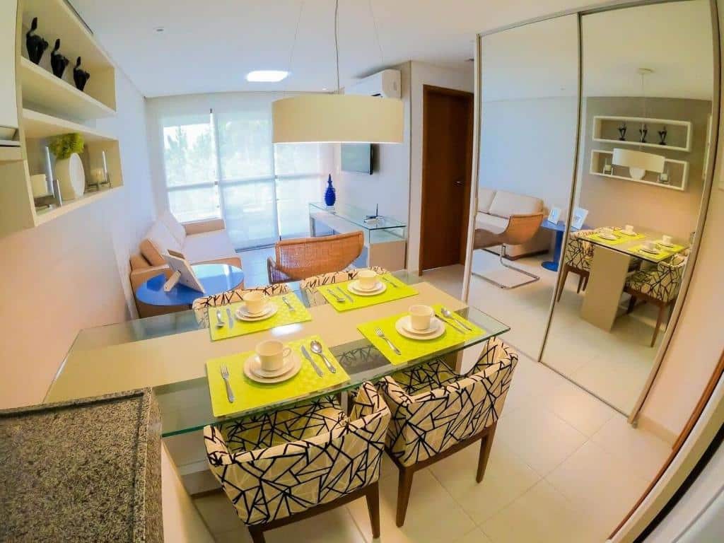 Interior de um apartamento do Carneiros Beach Resort. Há uma mesa com quatro cadeiras com talheres em cima, um ármário espelhado à direita e ao fundo uma sala de estar com sofá, ar-condicionado e TV suspensa. Há uma porta de vido com vista para árvores