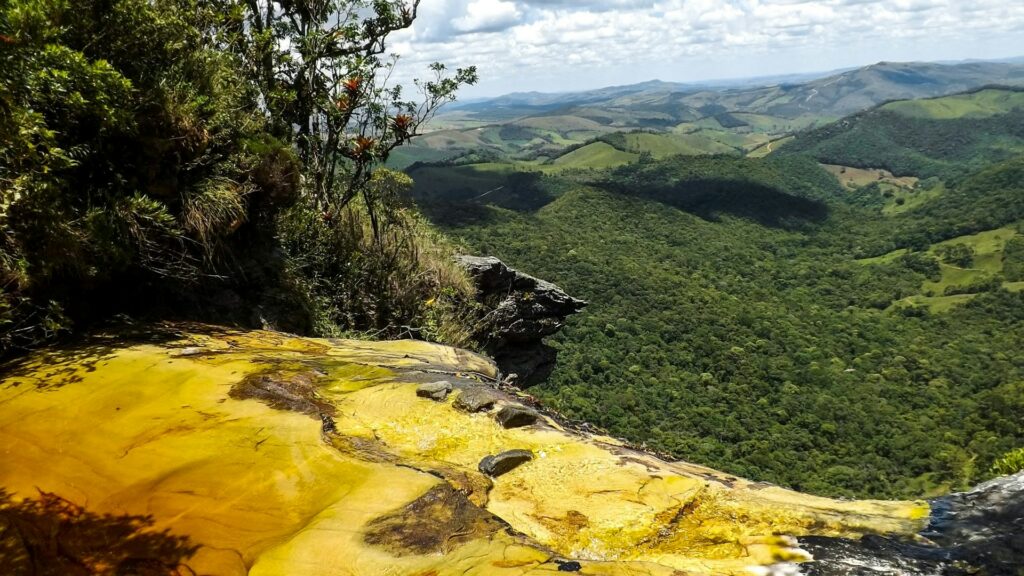 Rio de águas cor de coca-cola que deságua virando uma cachoeira em meio a árvores e plantações verdes e ao fundo é possível enxergar um vale verde de montanhas durante o dia. Imagem ilustrando post Airbnb em Ibitipoca.