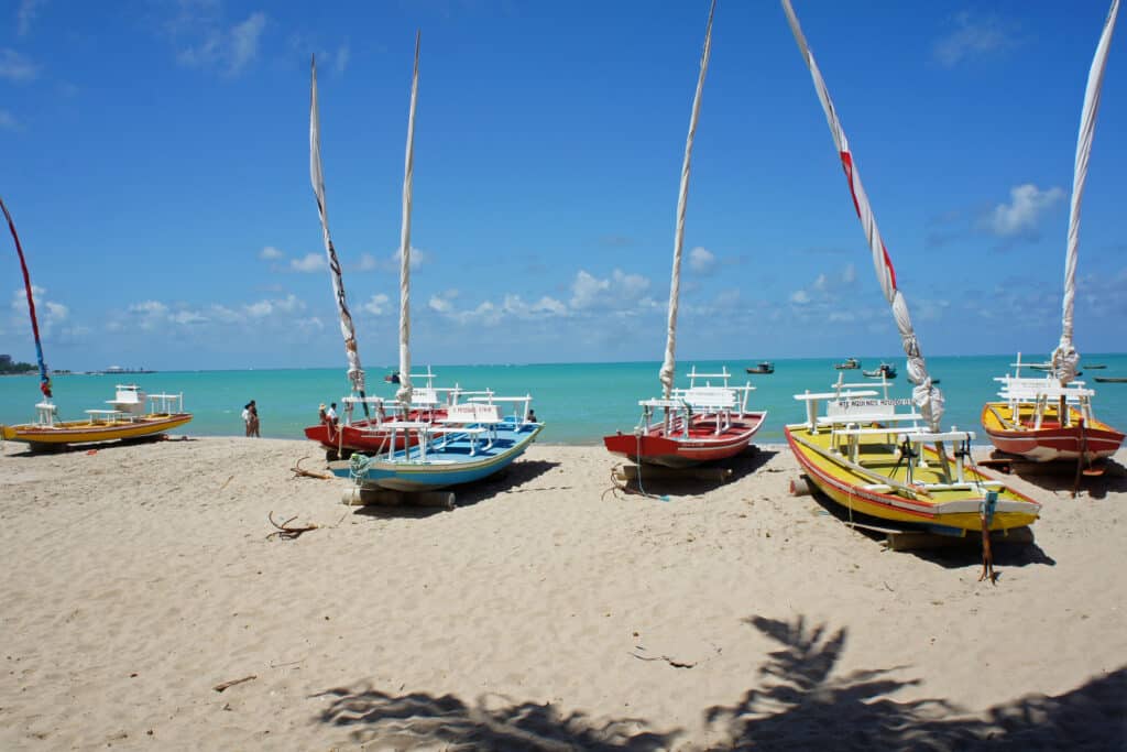 Jangadas de diferentes cores estacionadas na areia branca com um mar cristalino ao fundo. Há sombra de coqueiros à frente e a imagem foi tirada em um dia ensolarado.