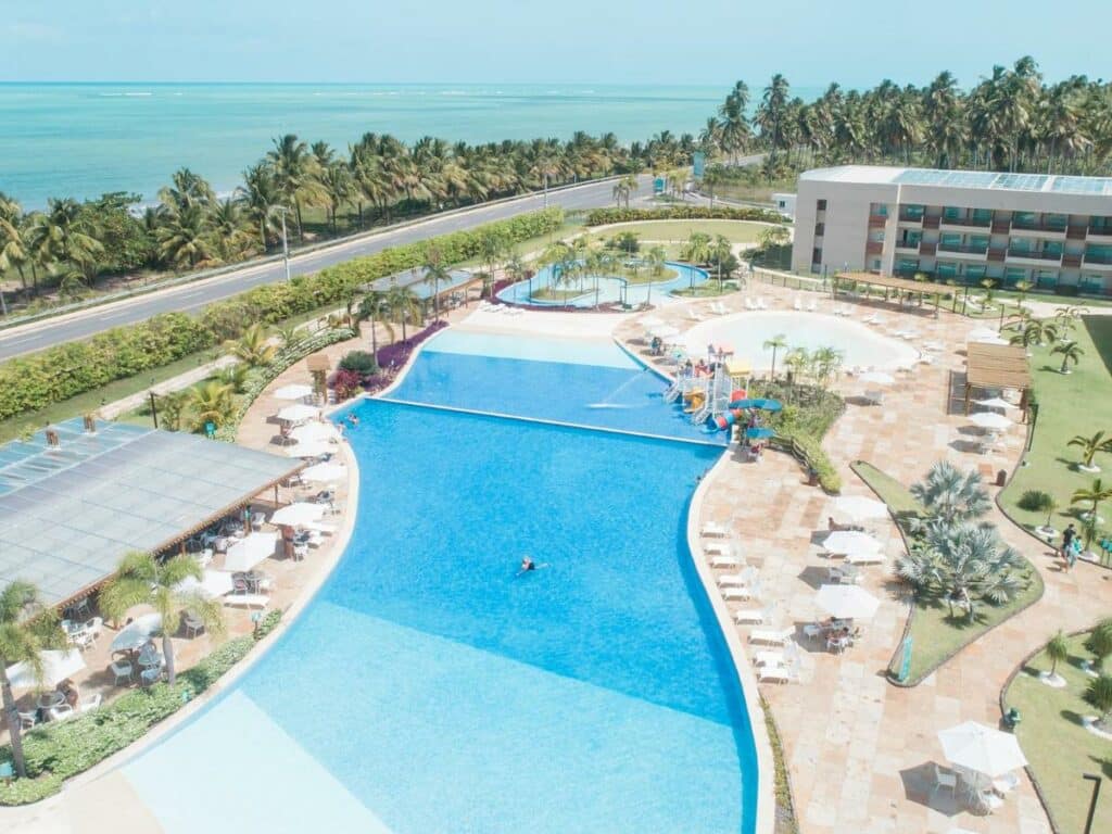 Foto da vista aérea do Japaratinga Lounge Resort. A imagem mostra uma vista aérea de um resort à beira-mar, com uma grande piscina azul que é o ponto central da fotografia. A piscina é de forma irregular e cercada por um deck com muitas espreguiçadeiras brancas e guarda-sóis. No canto direito da piscina, há uma área colorida com um pequeno parque aquático para crianças. O resort possui uma construção com telhado marrom e fachadas em tons claros. Além da área da piscina, há uma praia de areia branca com fileiras de coqueiros que separam o resort da costa. O mar é de um azul claro vibrante, e uma estrada corre paralelamente à praia. Representa resorts em Maragogi