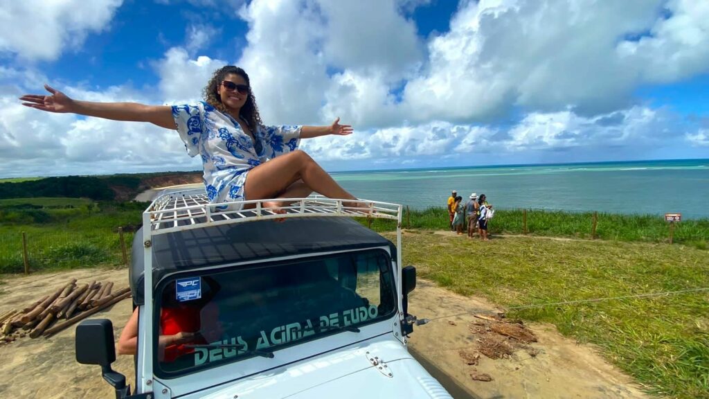 Mulher em cima de um JIpe com os braços abertos sorrindo. O carro está no mirante do Carro Quebrado, com gramado e vista panorâmica do mar.