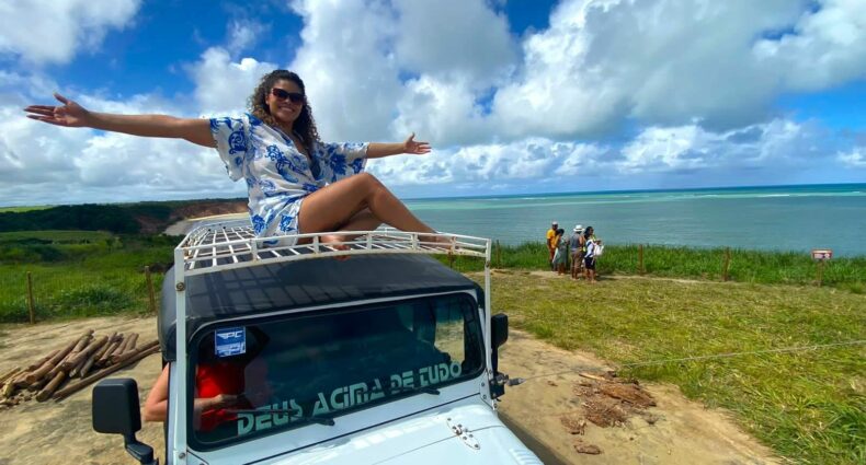 Mulher em cima de um JIpe com os braços abertos sorrindo. O carro está no mirante do Carro Quebrado, com gramado e vista panorâmica do mar. Representa o que fazer em Maceió e região
