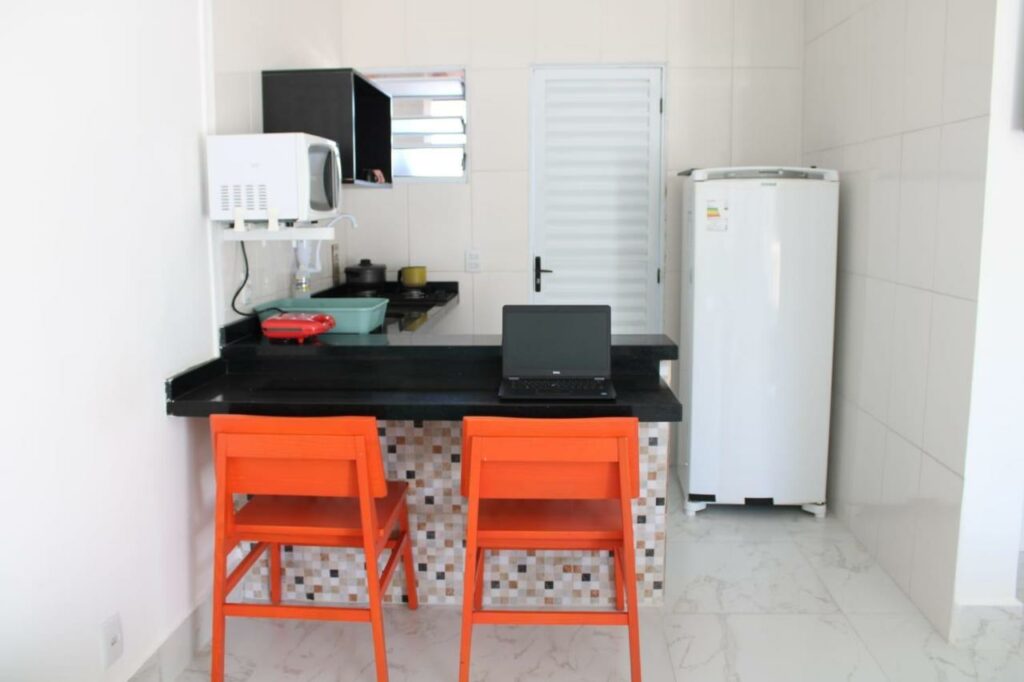 Cozinha da Kitnet Maitinga. No centro do ambiente há uma bancada com duas cadeiras e um computador. Ao fundo há utensílios ao lado da pia e um micro-ondas acima. Ao lado direito na cozinha fica a geladeira.
