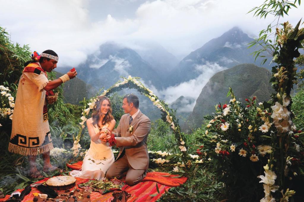 casal de noivos ajoelhados sobre uma manta vermelha, em meio a floresta peruana, com um xamã abençoando a união. Ao fundo é possível ver uma cadeira de montanhas, e ao redor dos noivos há várias flores decorando o ambiente.