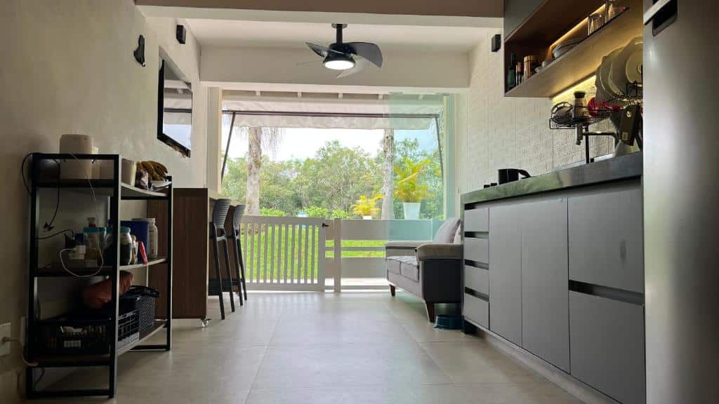 Cozinha do Lindo Loft em Condomínio Praia Barra do Una. Ao lado direito ficam armários com cooktop e utensílios de cozinha, além de uma geladeira, que encara uma prateleira na parede em frente. Ao fundo está a sala de estar com sofá ao lado das bancadas da cozinha e uma TV na parede em frente logo acima de uma mesa com dois banquinhos. Ao lado do sofá está uma porta com vista para fora.