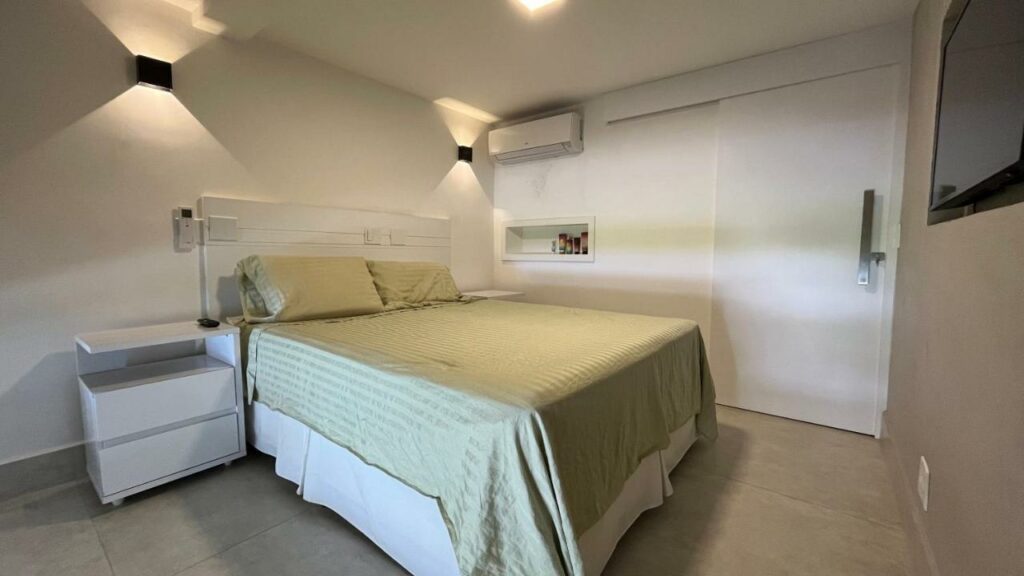 Quarto do Lindo Loft em Condomínio Praia Barra do Una, um dos airbnb em Barra do Una. Uma cama de casal tem mesinhas de cabeceira dos dois lados e encara uma TV na parede oposta. Um recorte na parede ao lado direito da cama tem amenidades de banho, e logo acima fica um aparelho de ar-condicionado.
