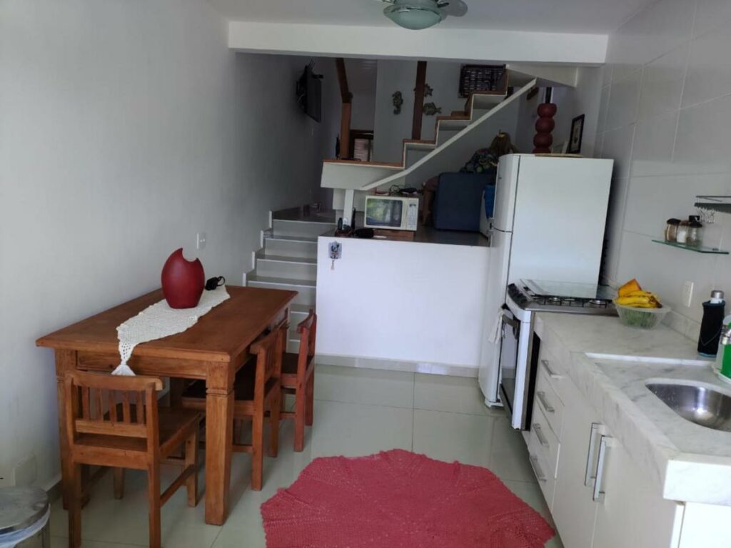 Cozinha do Lindo Loft na Praia de Barra do Una à beira do Rio. Um fogão está ao lado direito, entre uma bancada com pia e uma geladeira. Na parede oposta fica uma mesa de madeira com três cadeiras, e ao fundo está um micro-ondas ao lado das escadas que levam ao andar superior.