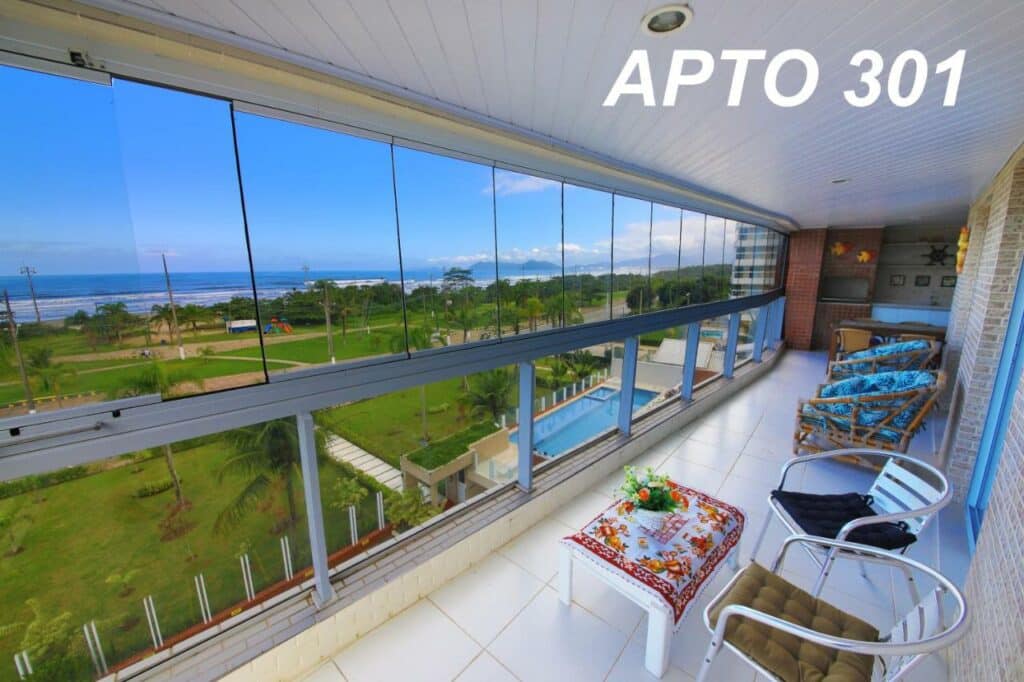 Vista do terraço do LINDO NOVISSIMO e PÉ NA AREIA. O local tem várias poltronas e cadeiras encarando o vidro com visão para uma área verde em frente à praia do Indaiá.