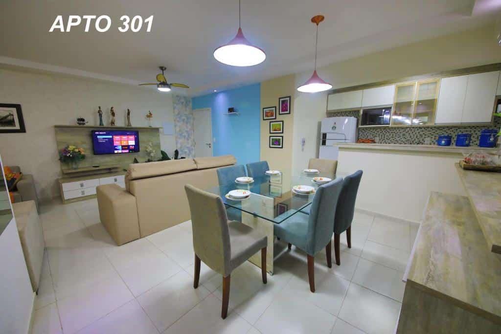 Visão geral do LINDO NOVISSIMO e PÉ NA AREIA. Ao lado direito da imagem fica uma mesa com seis cadeiras logo em frente à cozinha ao fundo, que tem geladeira, micro-ondas e armários atrás de uma bancada. Já do lado esquerdo fica a sala de estar, com sofás encarando um suporte com TV e ventilador de teto logo acima, para representar airbnb em Bertioga.