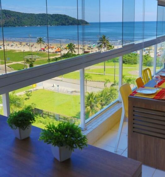 Vista do terraço do LINDO NOVISSIMO e PÉ NA AREIA, um dos airbnb em Bertioga. O local tem uma mesa com cadeiras e uma poltrona encarando o vidro com visão para uma área verde em frente à praia do Indaiá.