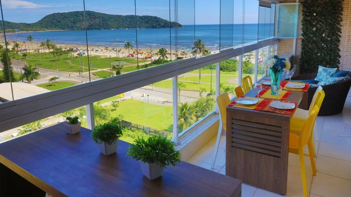 Vista do terraço do LINDO NOVISSIMO e PÉ NA AREIA, um dos airbnb em Bertioga. O local tem uma mesa com cadeiras e uma poltrona encarando o vidro com visão para uma área verde em frente à praia do Indaiá.