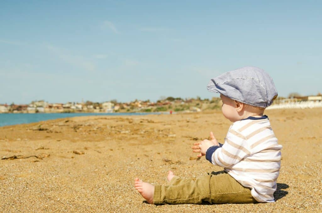 Um bebê sentado na areia de uma praia usando calça, blusa listrada e uma boina