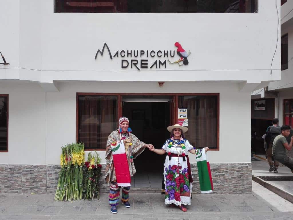 fachada de entrada do hotel machupicchu dream com um casal vestido com roupas temáticas do peru em pé na frente da porta e com as mãos dadas, olhando para a câmera. Eles estão sorrindo.