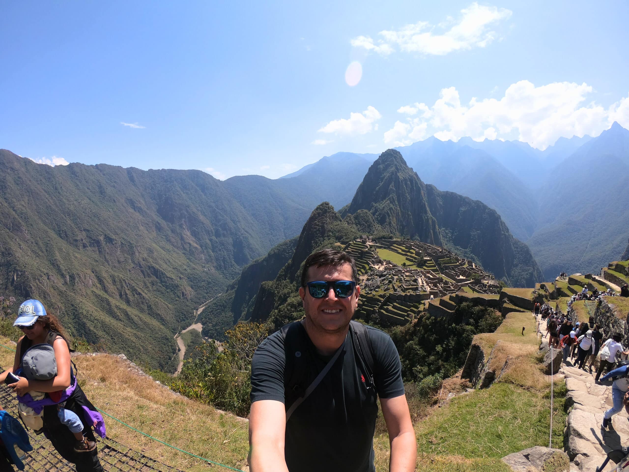 paisagem panorâmica de Machu Picchu, mostrando toda a cadeia de montanhas ao redor da cidadela, além do rio urubamba mais abaixo. No centro da imagem há um homem de óculos escuros e sorrindo para a câmera.
