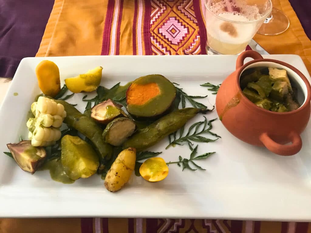 prato retangular branco com vários tipos de batatas e milhos, junto com uma cumbuca de barro com pães