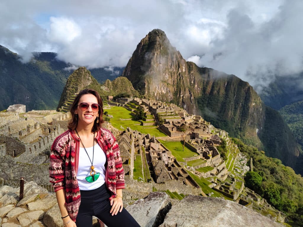 vista da cidadela de Machupicchu ao fundo, com um céu nublado e muita área verde. Próximo à câmera há uma mulher de cabelo curto, pele clara e óculos escuros sorrindo. Ela usa um casaco vermelho com desenhos incas e um colar comprido, preto e verde.