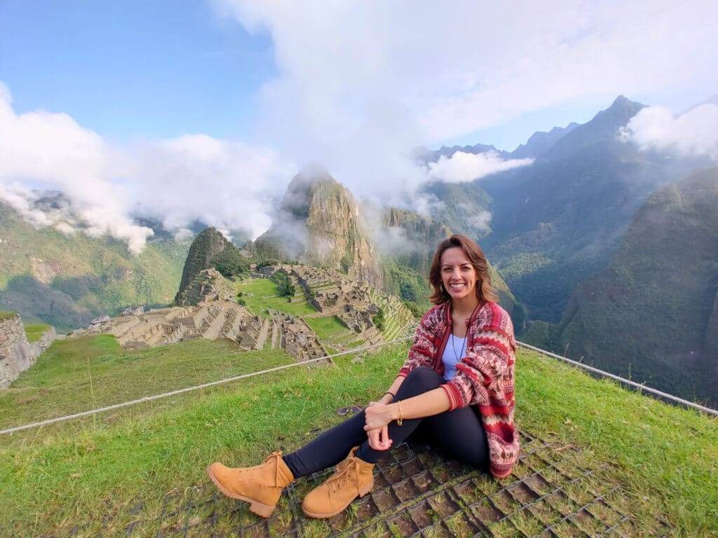 nátalie guimarães sentada no gramado, sorrindo para a câmera, com a paisagem de fundo mostrando a cidadela e as montanhas incas de Machu Picchu