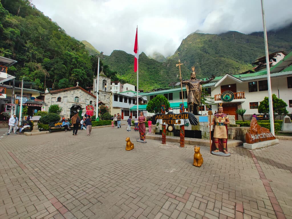 vista da Plaza Manco Capac em Machupicchu pueblo msotrand a igreja ao fundo e uma estatueta indígena à frente, no lado direito da imagem. Mais atrás é possível ver a cadeia de montanhas que cerca a cidade, assim como as nuvens carregadas de chuva que as sobrevoam