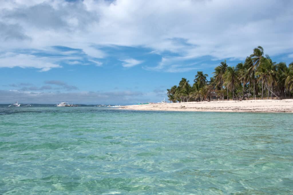 Ilha de Malapascua, em Cebu, com água turquesa clara, areia branca e palmeiras sob um céu azul parcialmente nublado. Barcos estão à distância.
