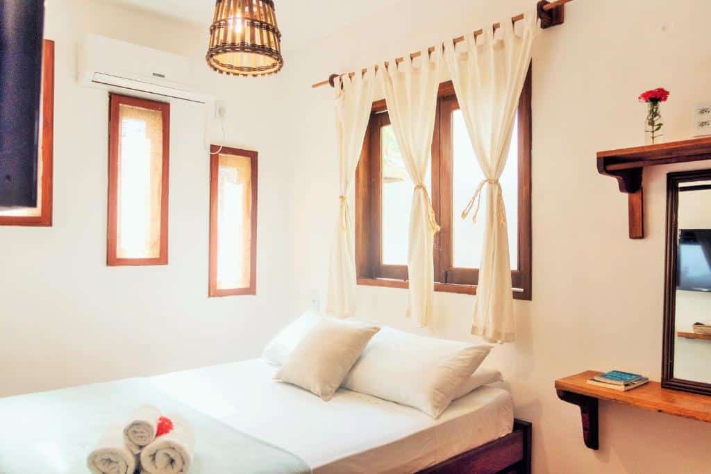 Foto do quarto do Manay Flats. Representa o post sobre airbnb em Jericoacoara. Há uma cama de casal. Atrás dela há uma janela. Ao seu lado, na direita, há duas prateleiras e um espelho pequeno. Na esquerda, hámais três janelas verticais e um ar-condicionado.