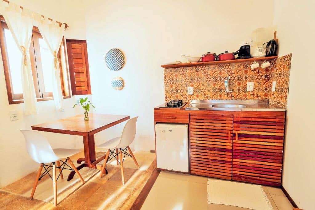 Cozinha compacta do Manay Flats. Uma pia com armário, frigobar e utensílios para cozinhar está na esquerda. Na direita há uma mesa com duas cadeiras e uma janela ao lado.