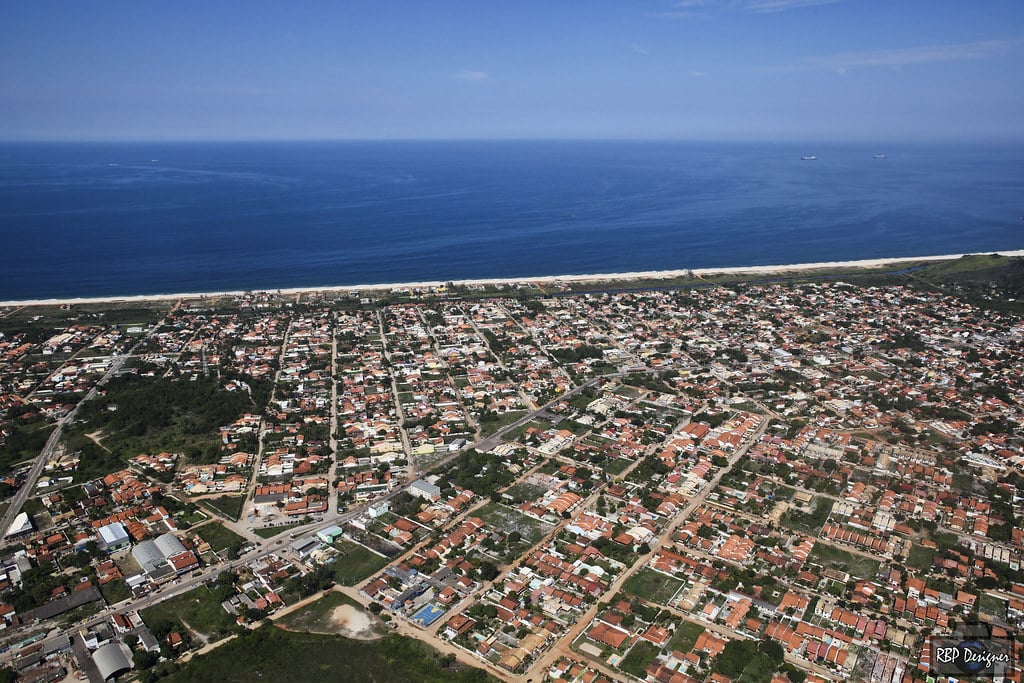 Vista de cima de Maricá, uma cidade grande na frente e atrás um mar azul com um céu sem nuvens.