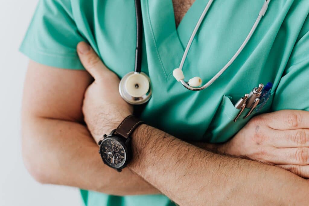 close nos braços cruzados de um médico com relógio, estetoscópio no pescoço e roupa privativa verde