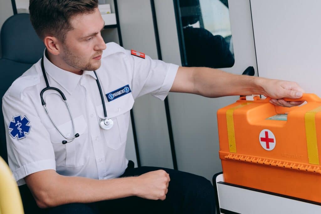 homem médico com maleta com estetoscópio no pescoço sentado, segurando uma maleta de primeiros socorros laranja dentro de uma ambulância