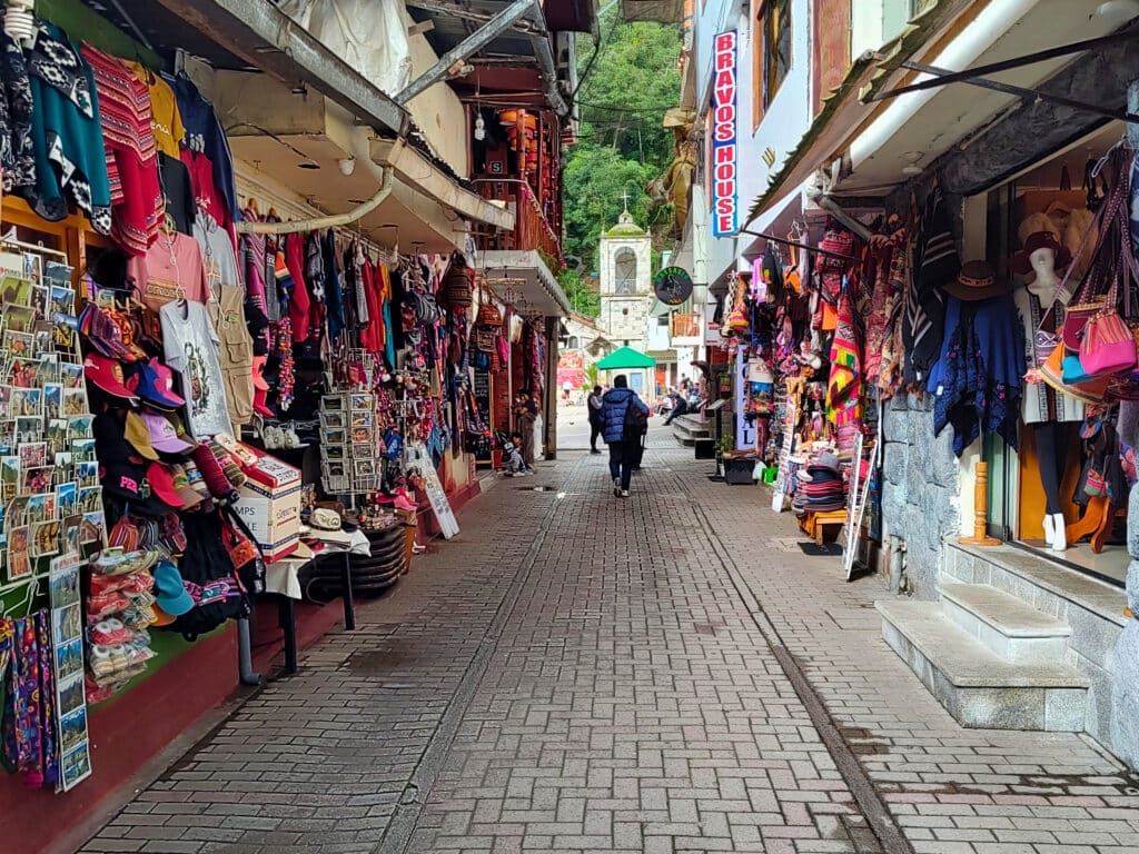 ruas de Machupicchu Pueblo com os dois lados repletos de produtos artesanais, coloridos e variados. Ao final da rua é possível ver a torre de sino da igreja na praça central.
