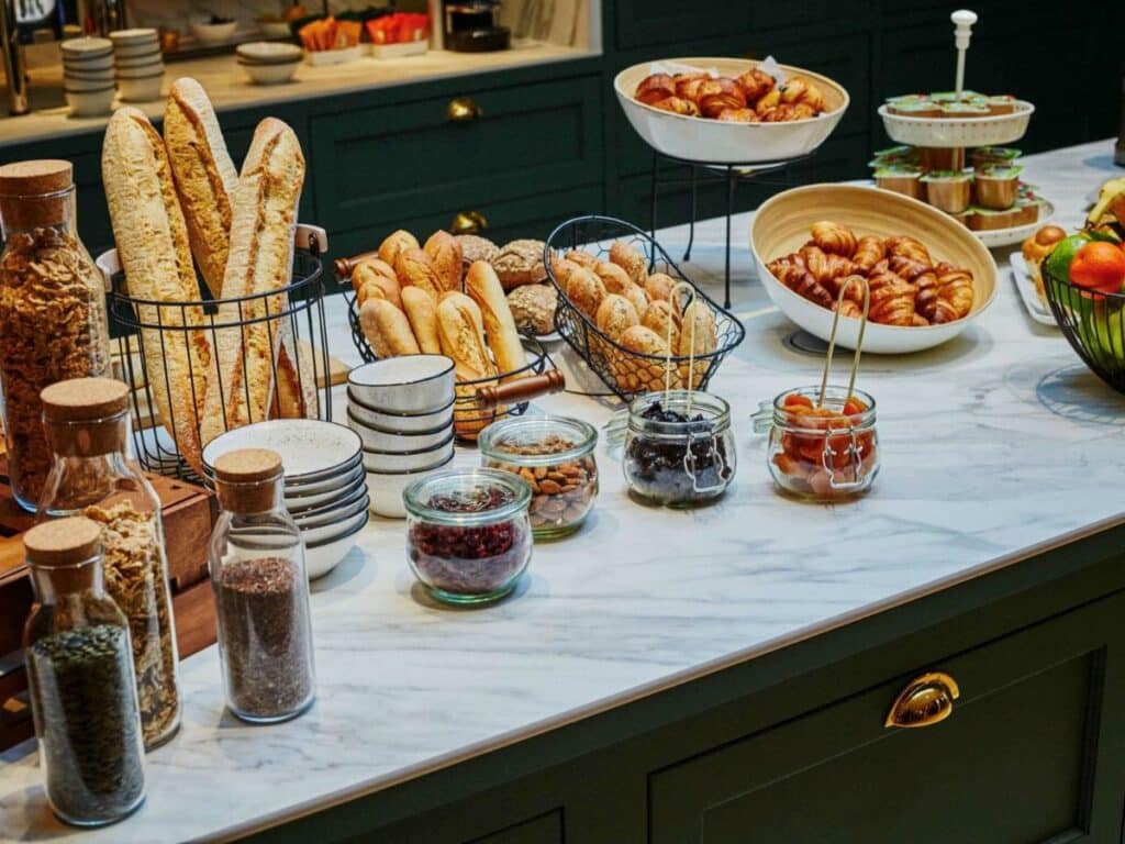 Balcão com itens de café da manhã no Mercure Versailles Chateau. Pães, cereais e frutas são alguns dos alimentos dispostos ali, e ao fundo está outro balcão com tigelas e utensílios.