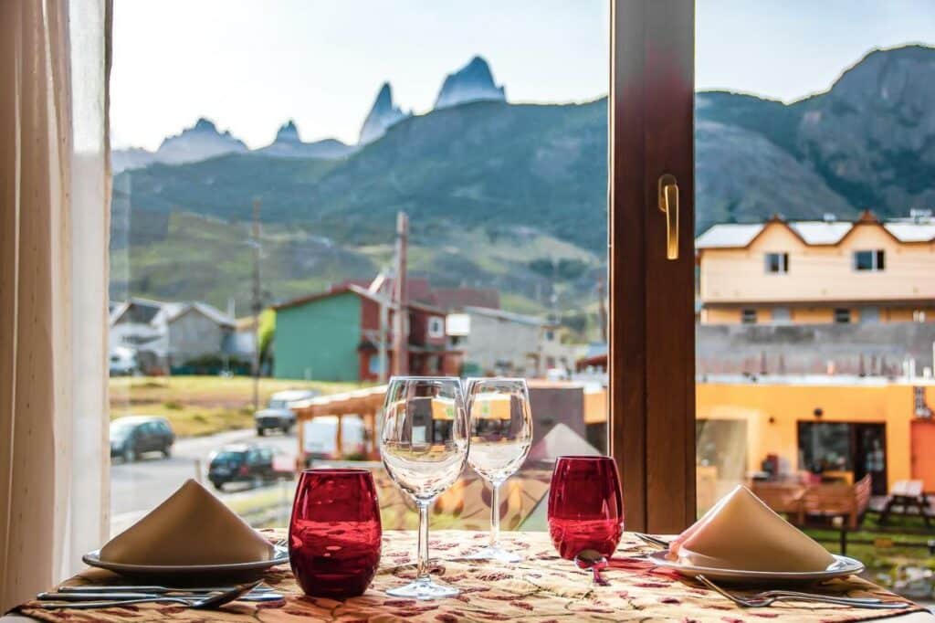 Mesa de jantar do Chalten Suites Hotel durante o dia com a mesa a frente com duas taças e dois copos com uma janela ao fundo com vista da cidade e montanhas.