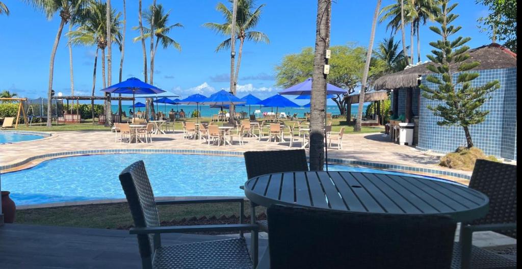 Mesa redonda com cadeiras em volta com a vista ao fundo de uma piscina, mesas com guarda-sóis e o mar ao fundo. Há árvores espalhadas e o céu está ensolarado