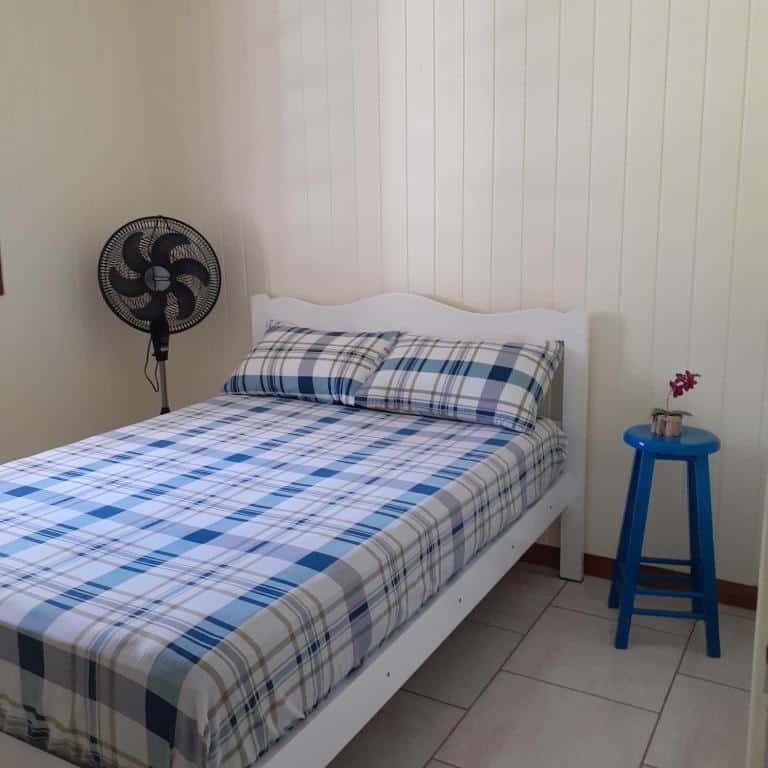 Foto do quarto em Meu pedacinho de chão. Há uma cama de casal, e na sua esquerda há um ventilador enquanto na direita há um pequeno banco pintado de azul com uma flor em cima.
Ilustra o post sobre airbnb em Serra Negra.